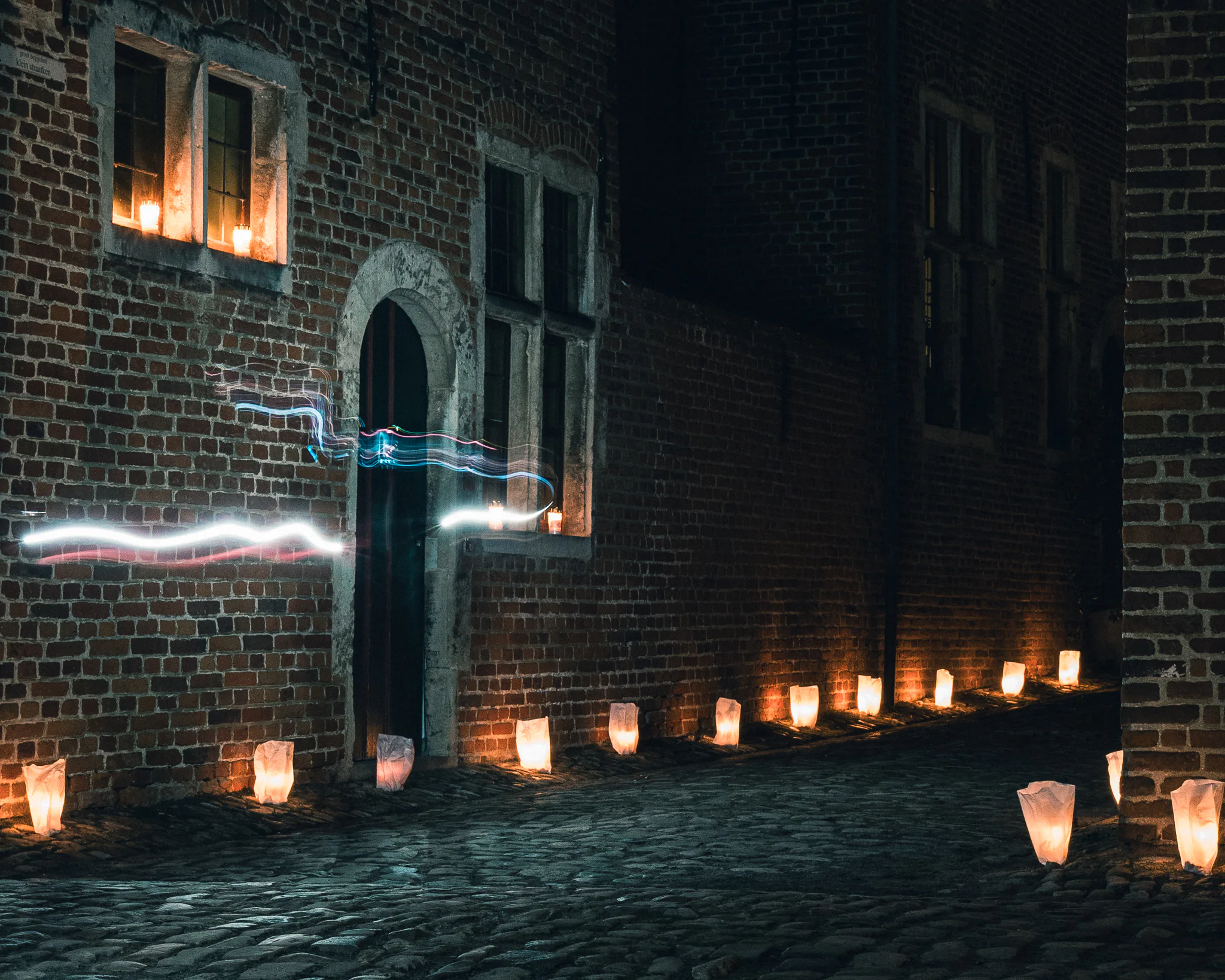 A candlelit brick lane crossed by a soft trail of moving light.