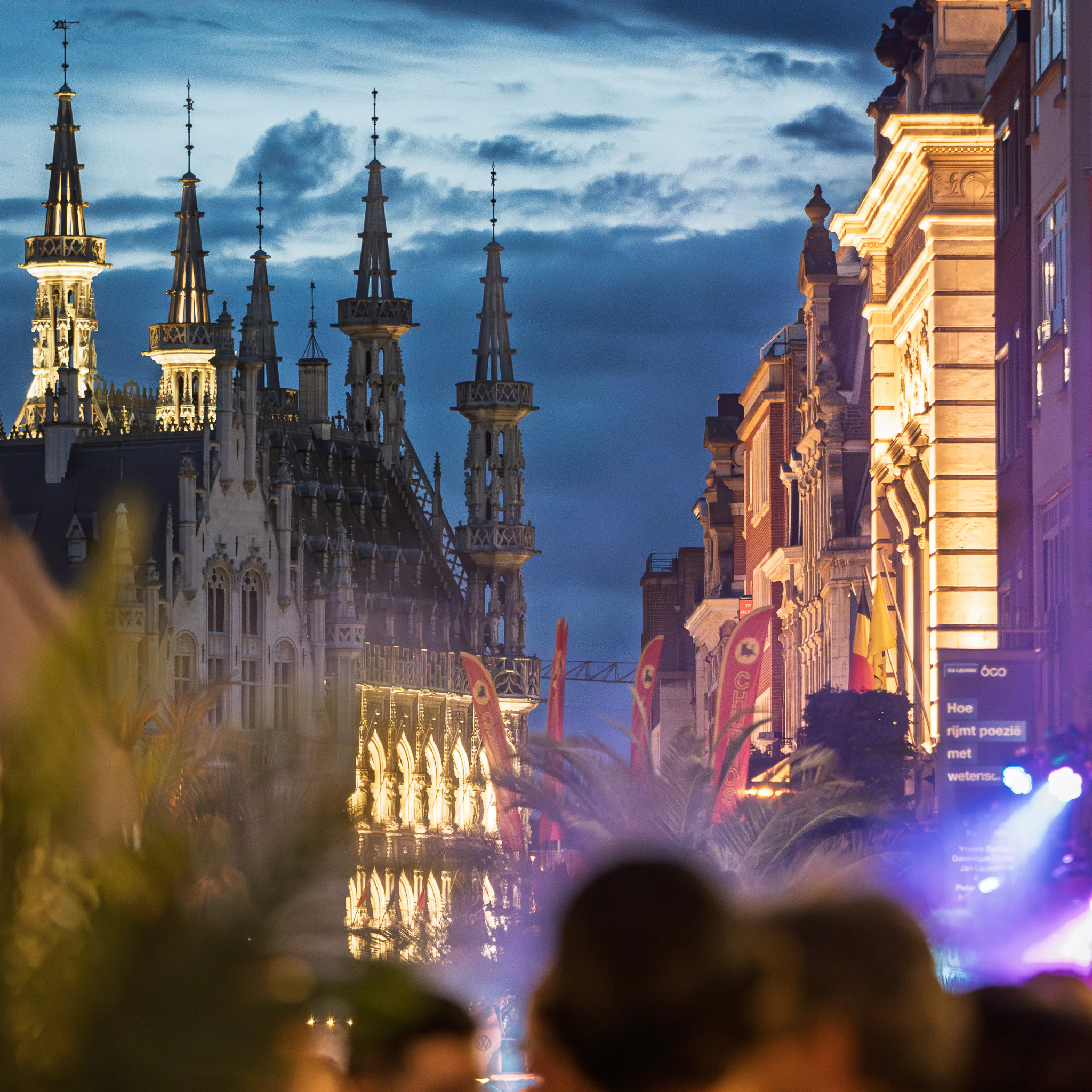 Leuven’s illuminated towers seen through a festival crowd and hazy stage light.