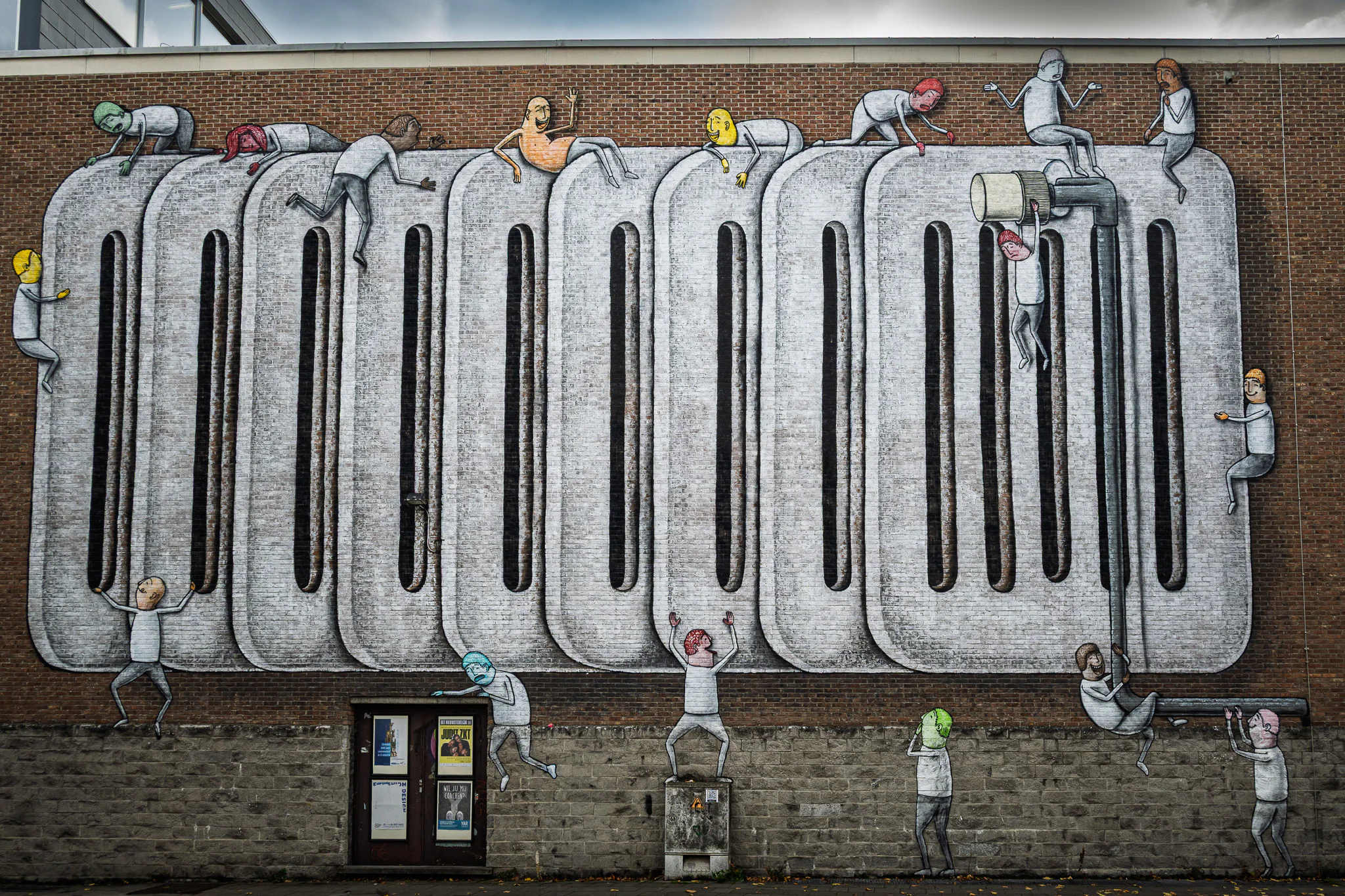 A long brick wall covered by a mural of small figures and monumental grey forms.