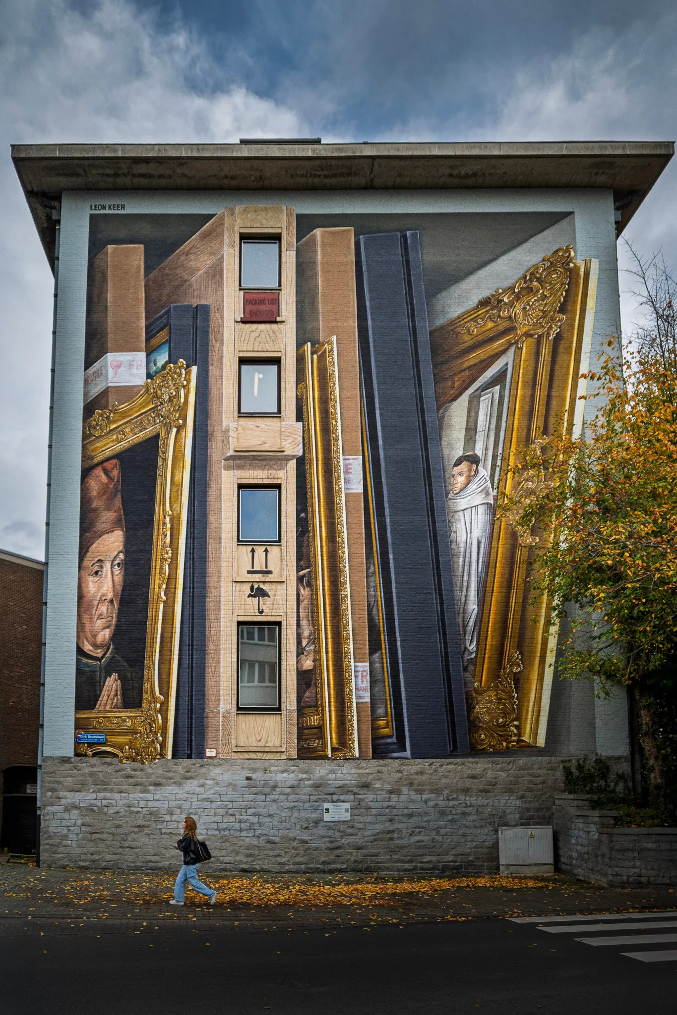 A pedestrian walking beneath a full-height mural of stacked gilded picture frames and painted portraits on a Leuven building.