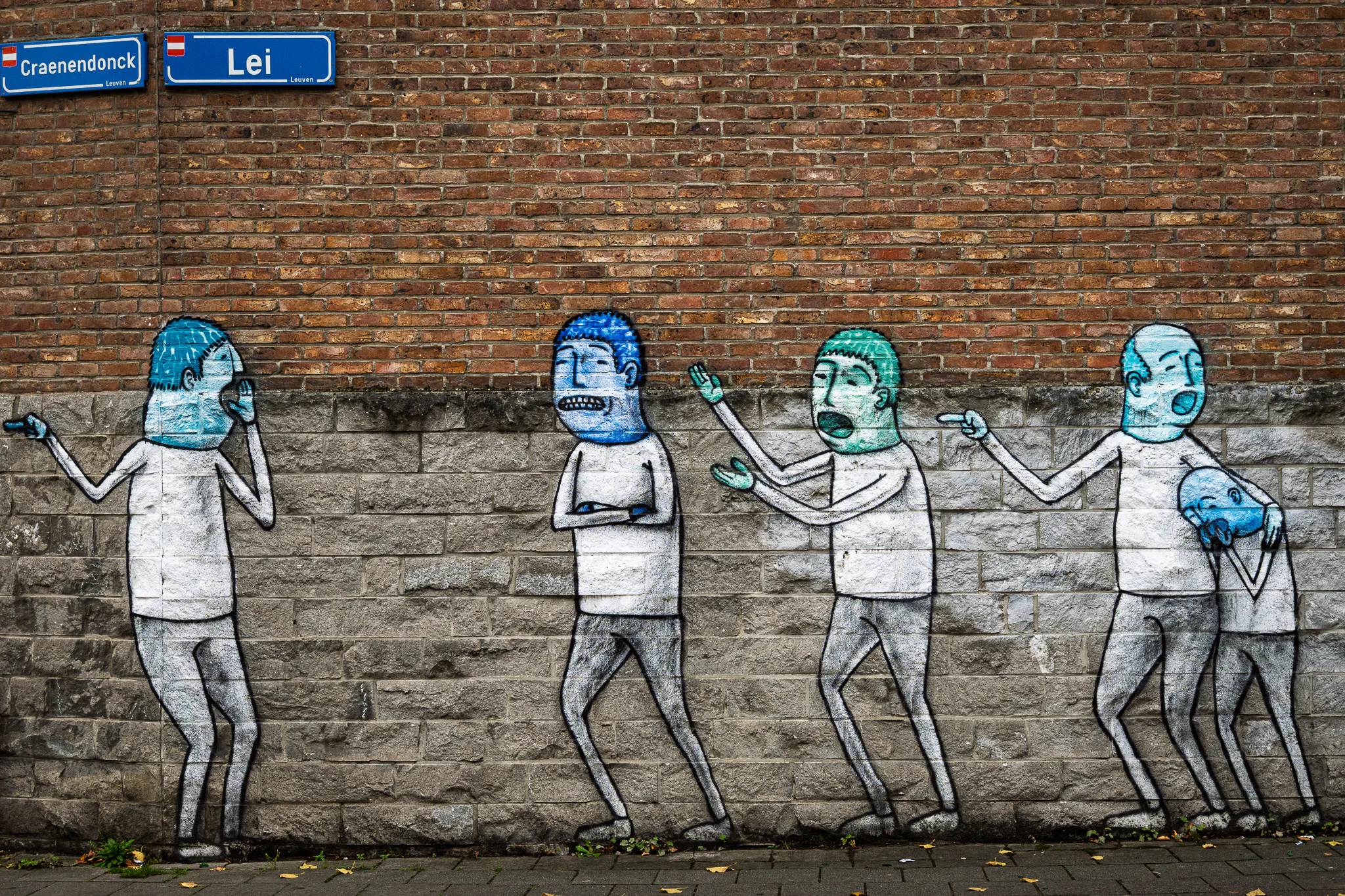 Five painted figures gesture and call to one another along a brick wall beneath Leuven street signs.