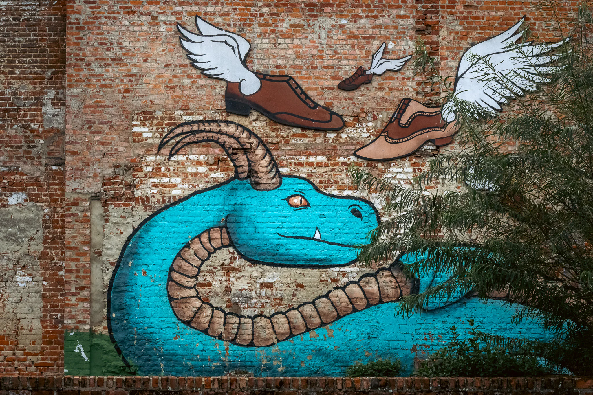 A turquoise dragon mural with flying winged shoes on an old brick wall, partly crossed by tree branches.