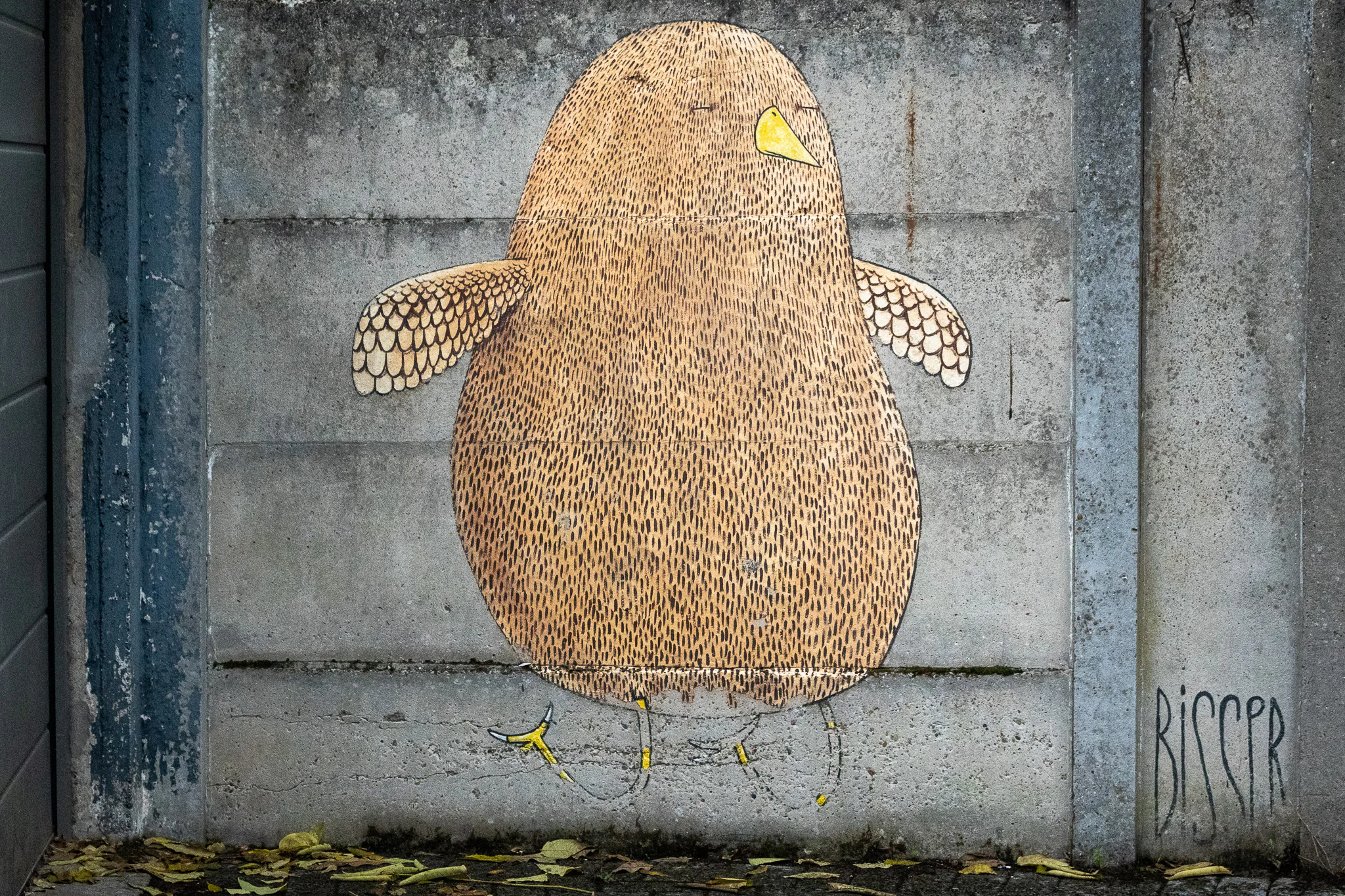 A rounded ochre bird painted on grey concrete as part of Bisser's Blue Cat and birds mural.