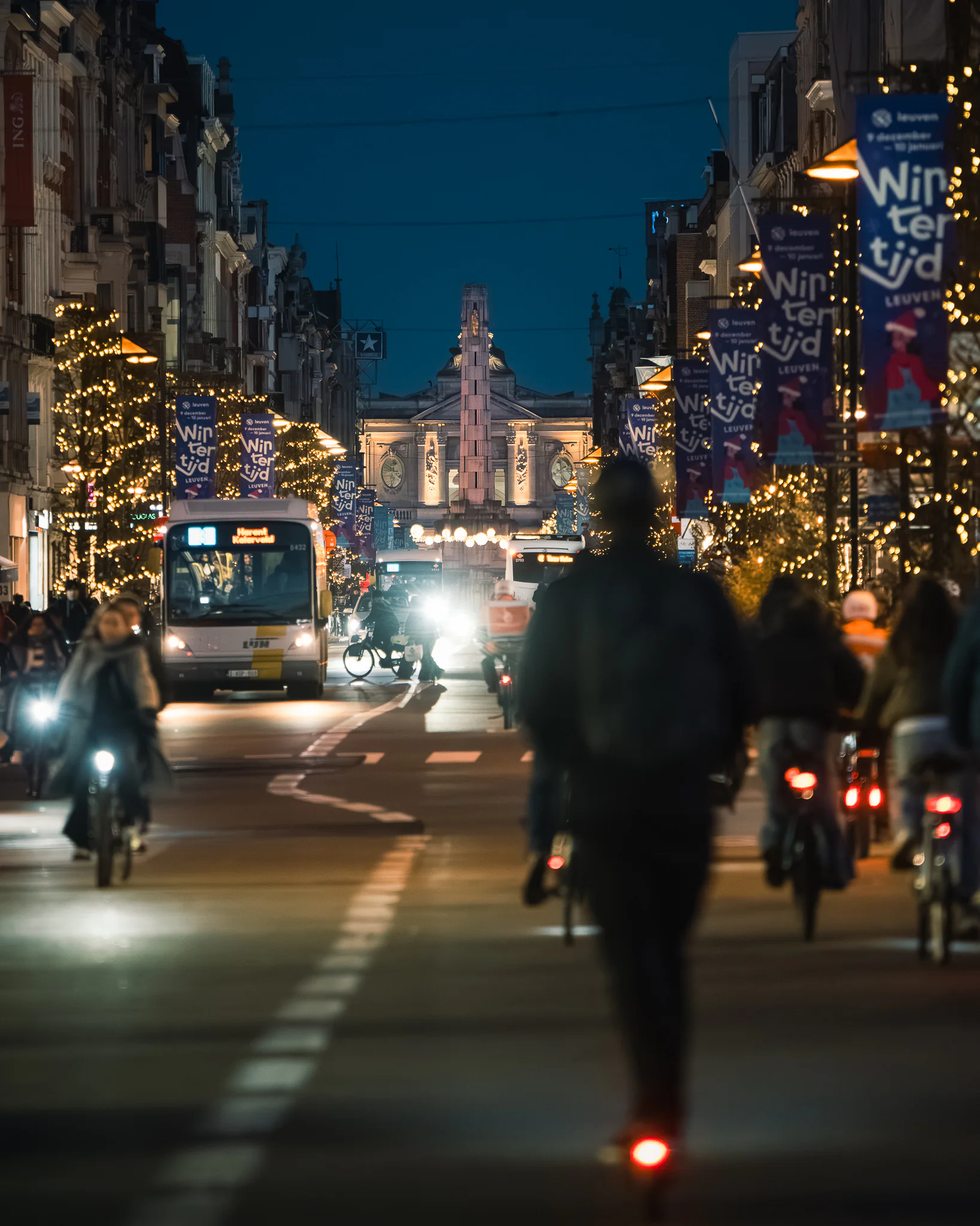 Cyclists and traffic moving along an illuminated Leuven street on a winter evening.