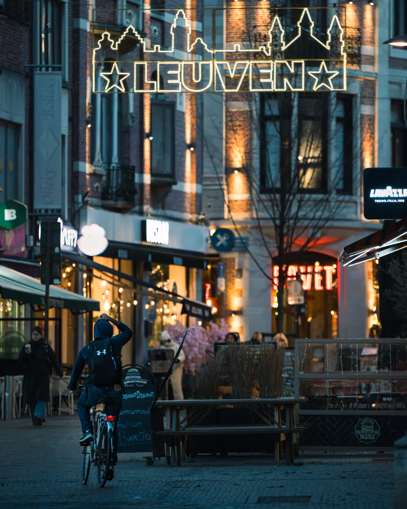A cyclist passing beneath an illuminated Leuven sign on a winter evening.