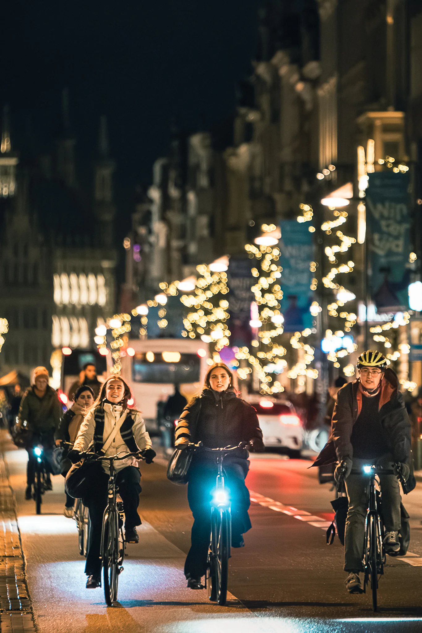 Two cyclists riding toward dense seasonal lights on a busy city street.