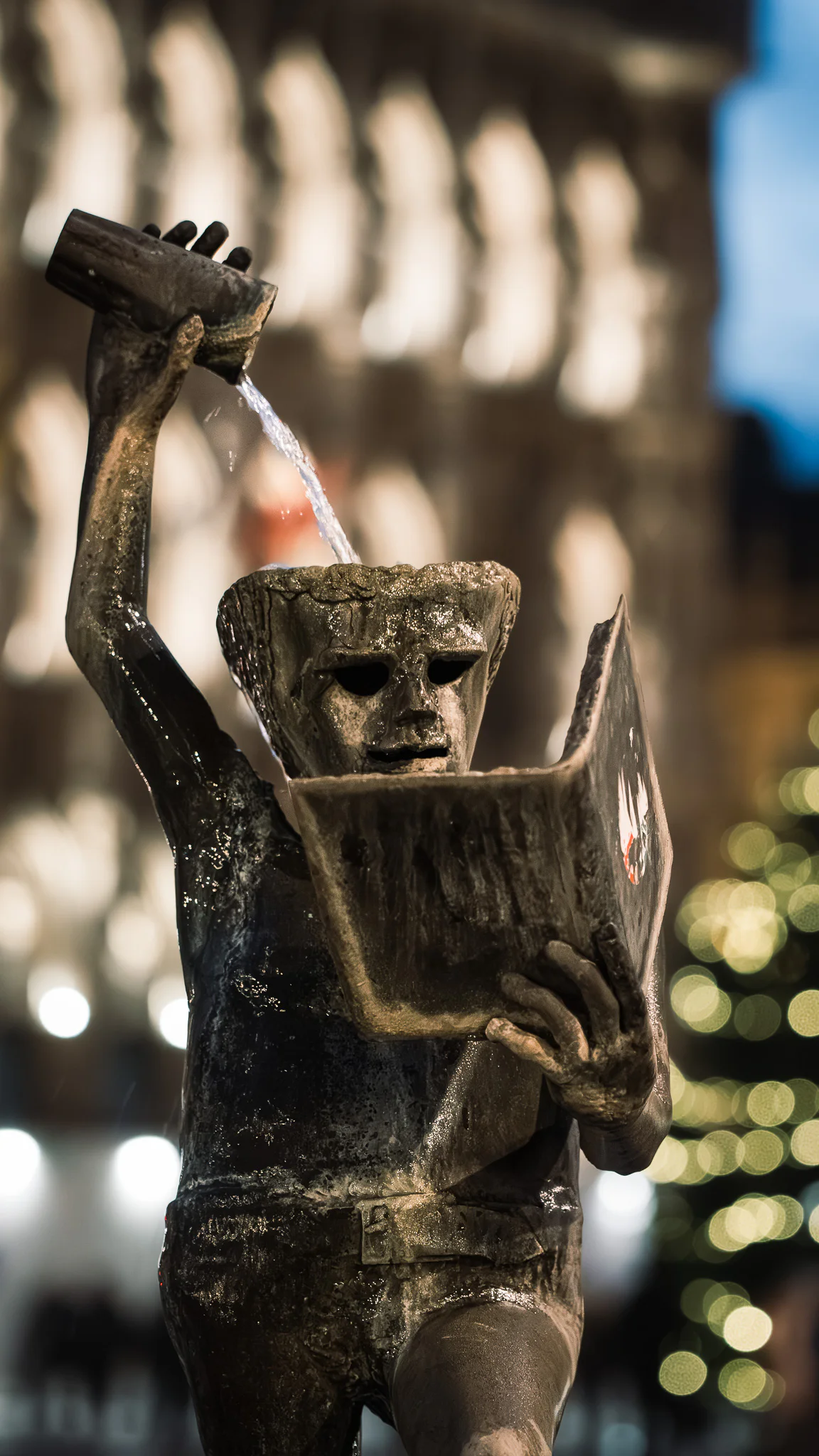The bronze Fons Sapientiae fountain figure seen against soft winter lights after rain.