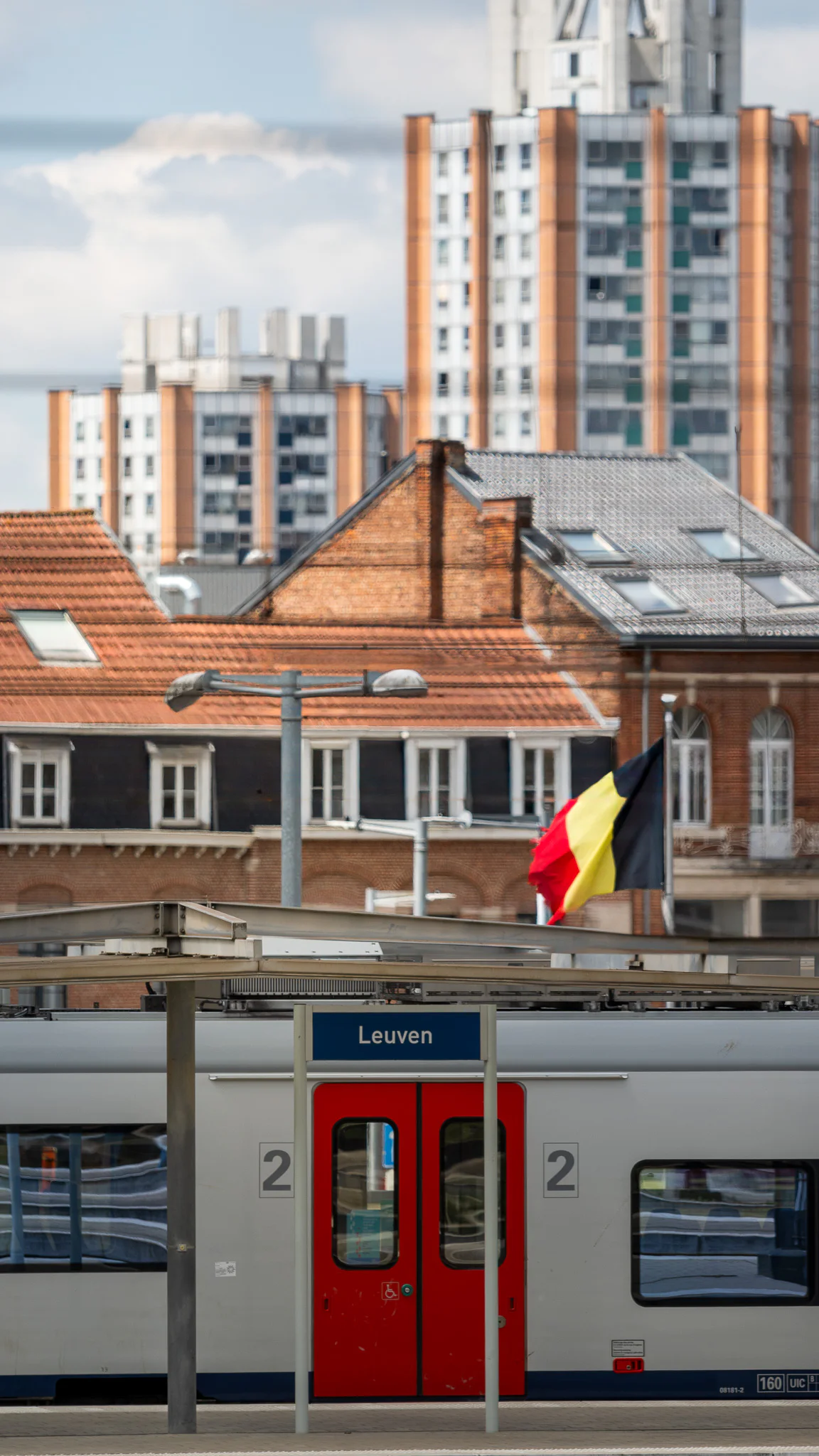 An NMBS train at Leuven station beneath a Belgian flag, with brick rooftops and the Sint-Maartensdal housing towers behind it.