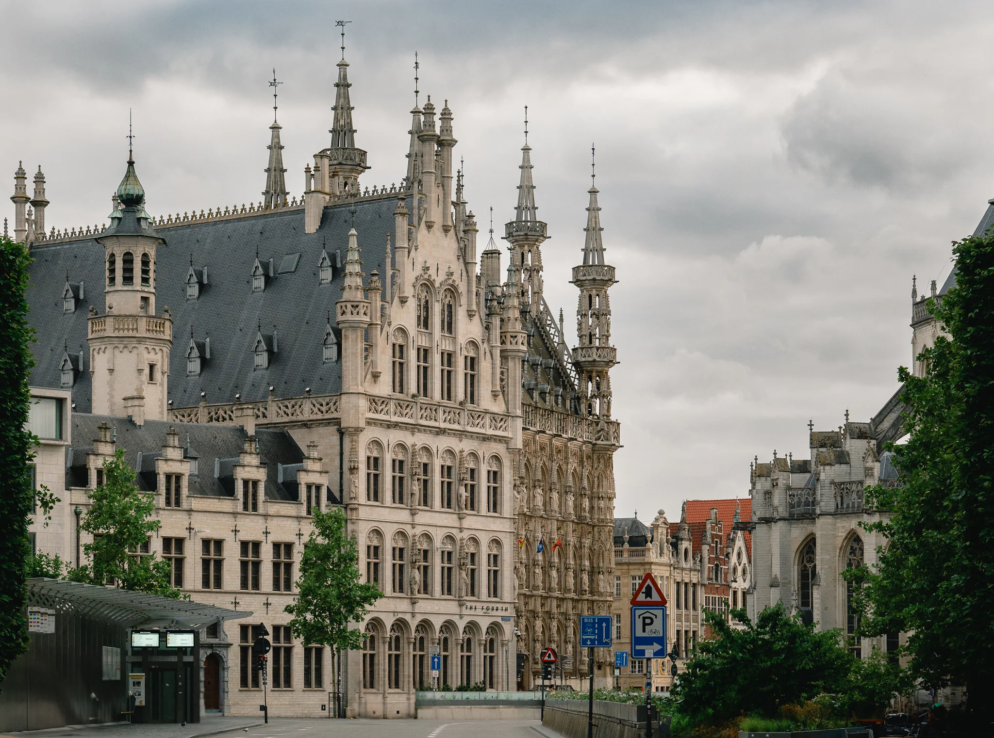 Leuven Town Hall and the edge of Saint Peter's Church beneath heavy storm clouds, framed by trees and city buildings.