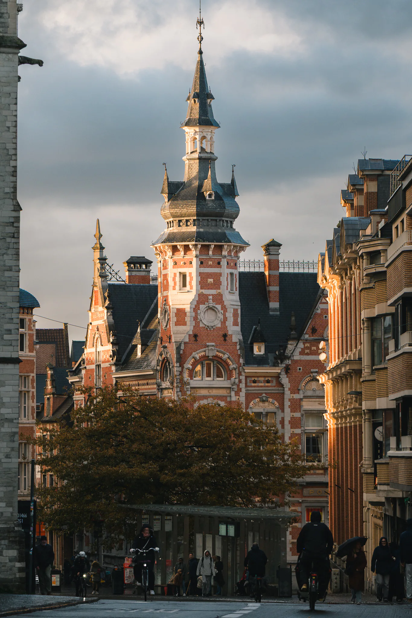 The red-brick tower and ornate facade of Leuven's former Main Post Office catching warm evening light above pedestrians and cyclists.