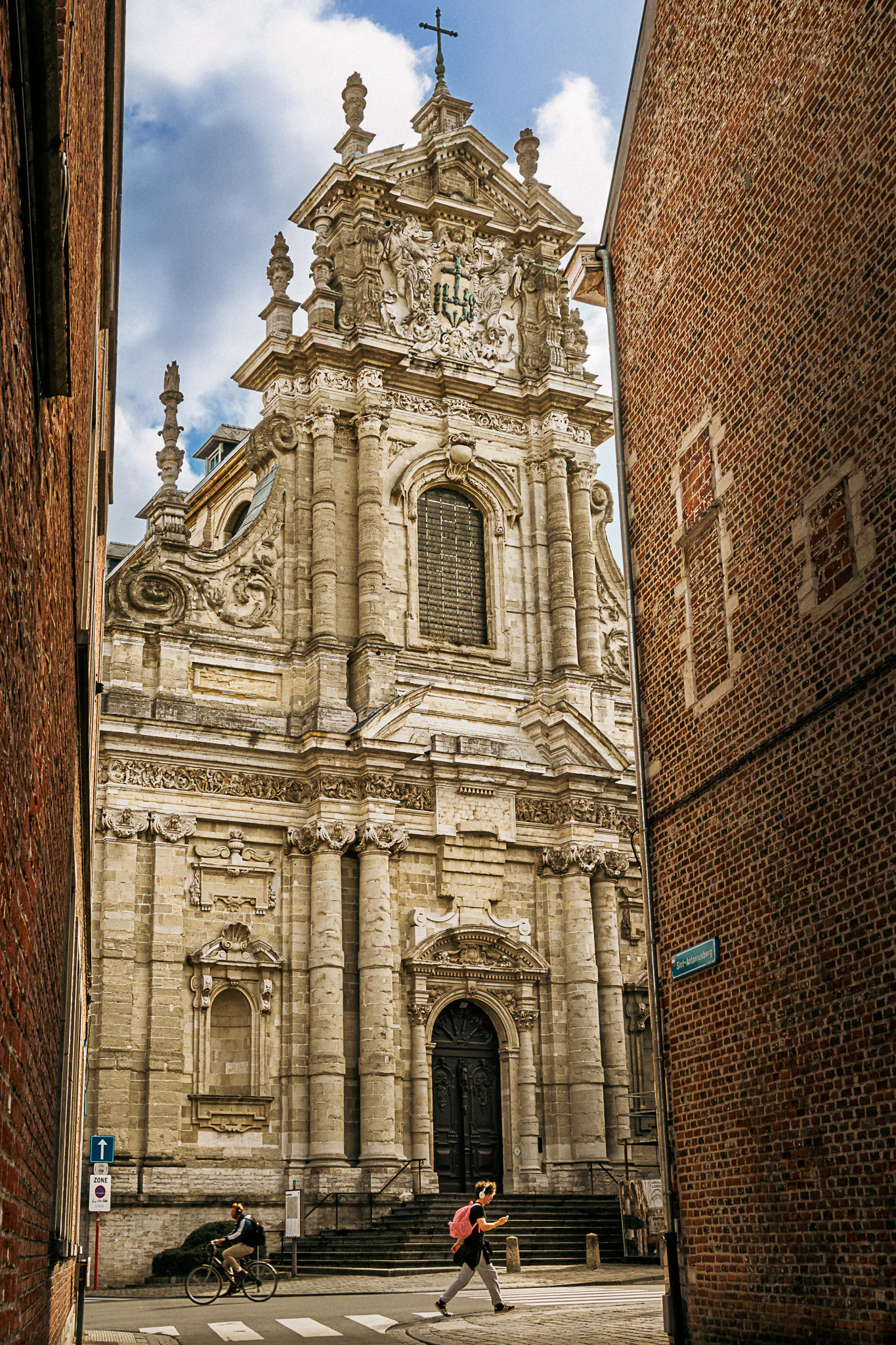 A pedestrian and cyclist crossing below the ornate Baroque facade of Saint Michael's Church, framed by narrow brick walls.
