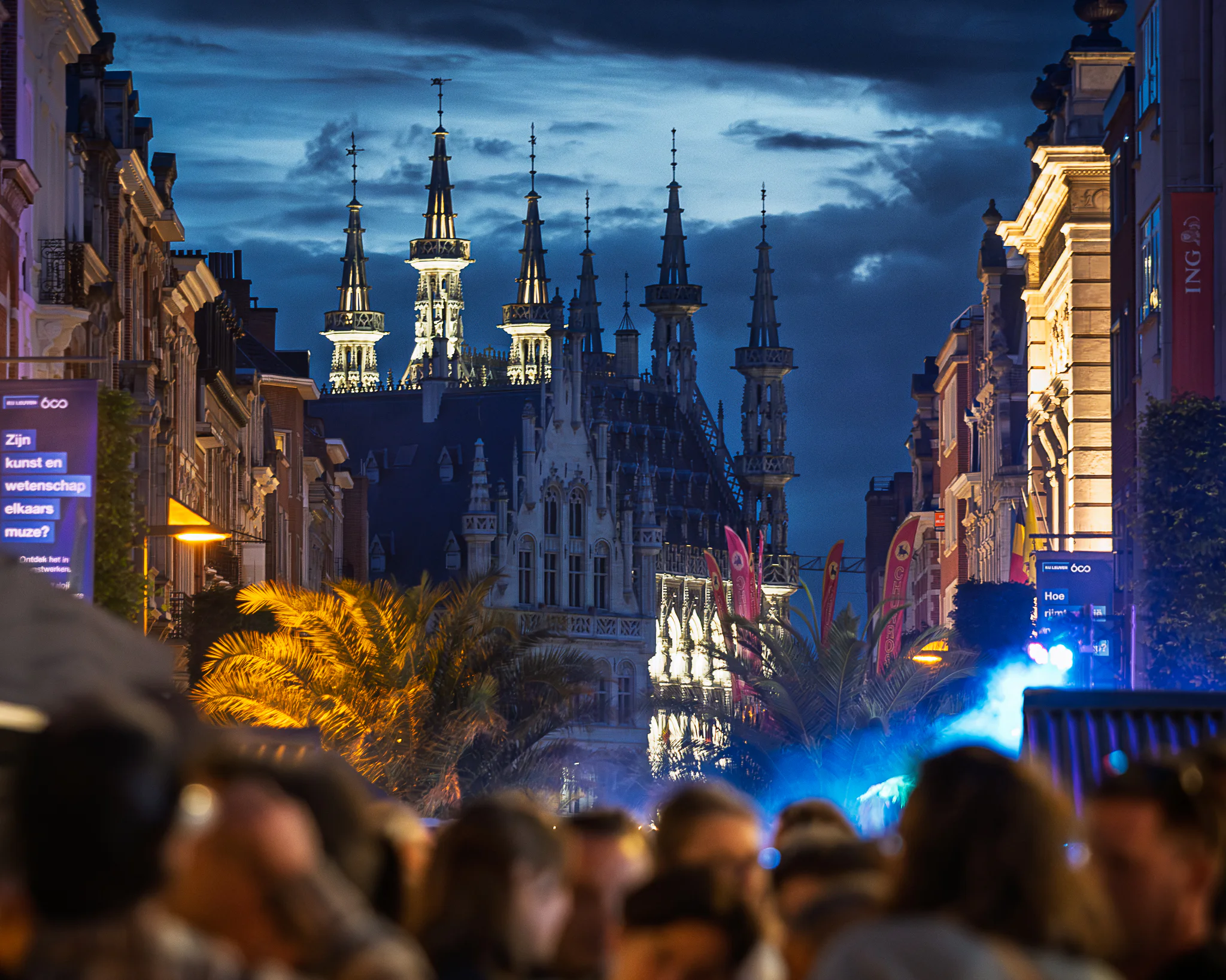 Leuven Town Hall illuminated at blue hour beyond a dense crowd, temporary palm trees, banners and blue event light.