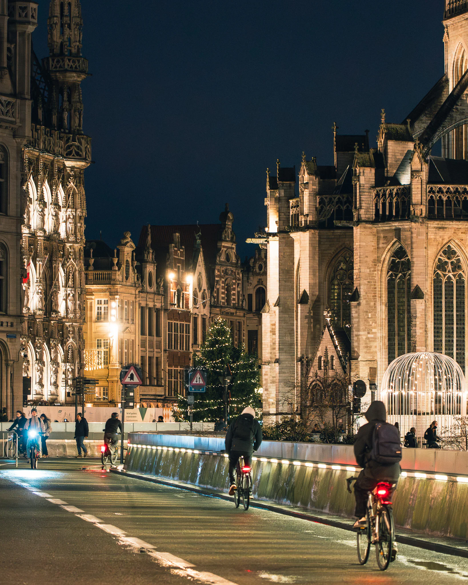 Cyclists riding along a wet street at night between illuminated Leuven Town Hall and Saint Peter's Church.